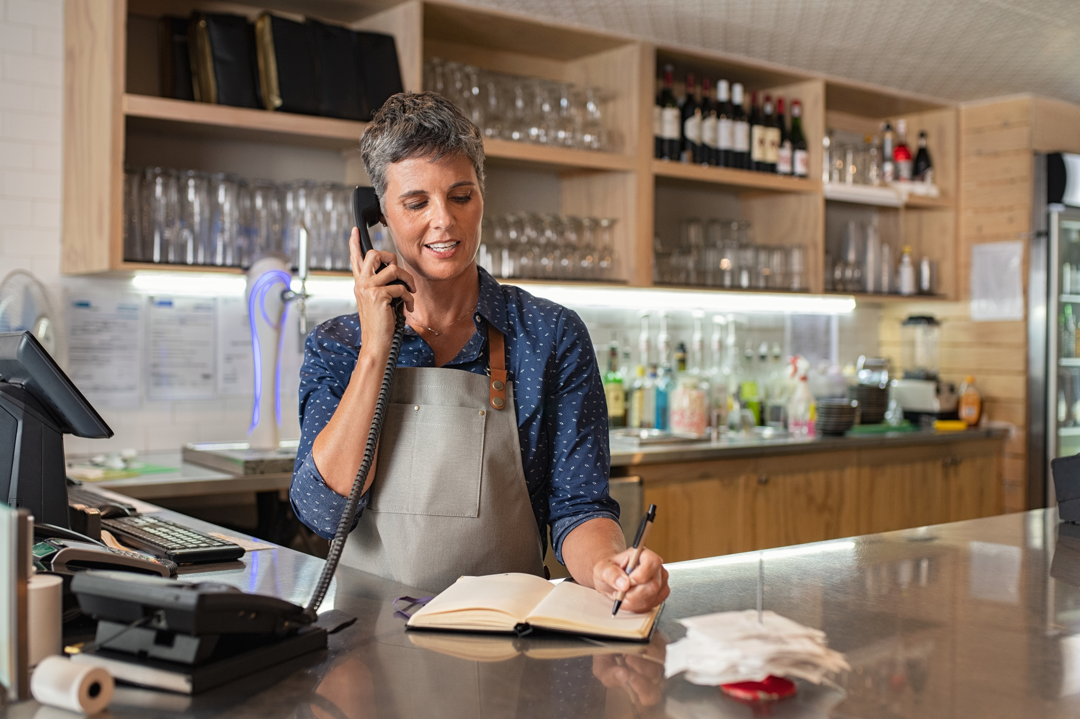 Barista woman taking orders over the phone at counter. Mature woman in grey apron writing on customer book while talking over phone in cafeteria. Waitress writing takeaway lunch while using telephone.